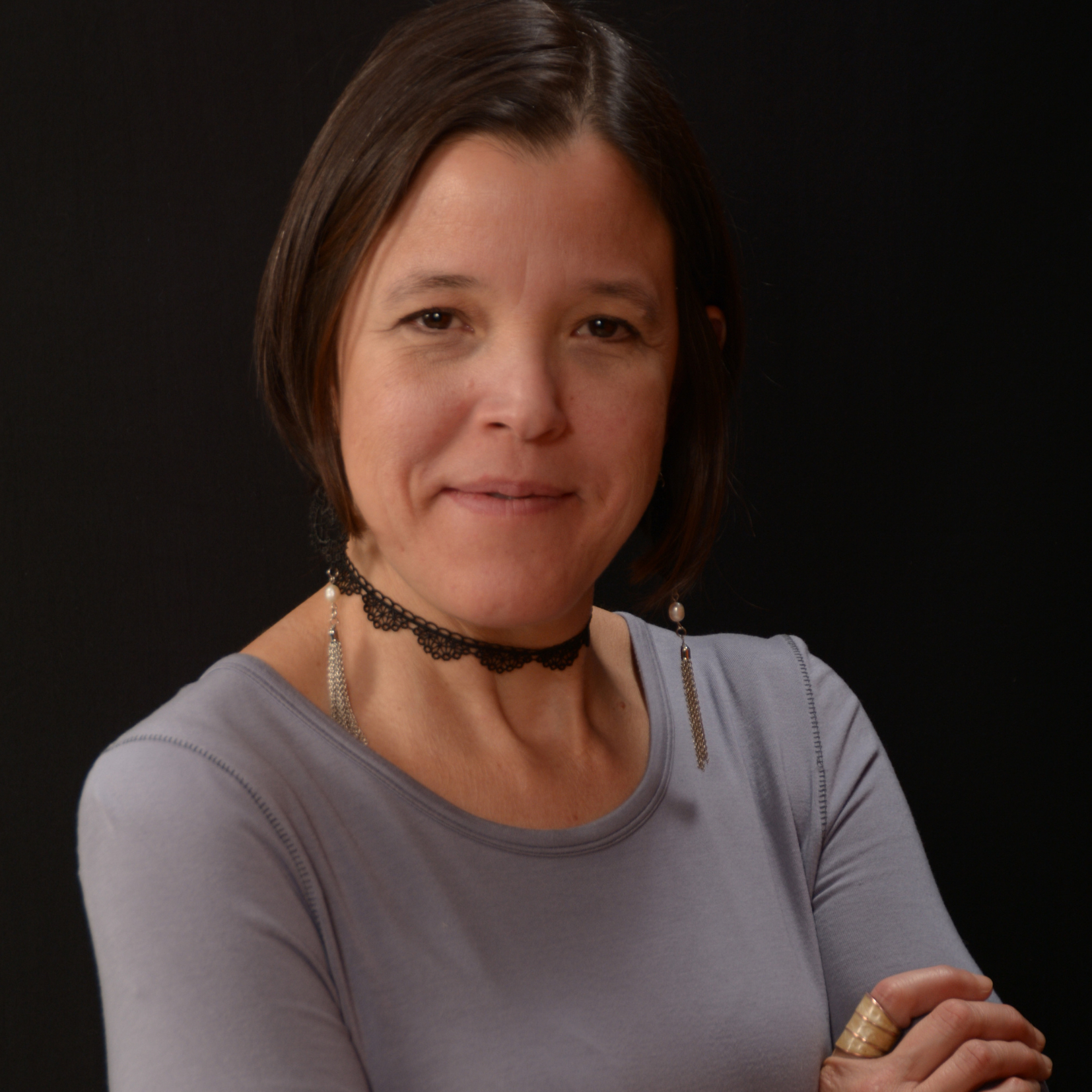 A woman with short brown hair, wearing a gray top and a necklace adorned with fantasmitas, poses with arms crossed in front of a dark background.