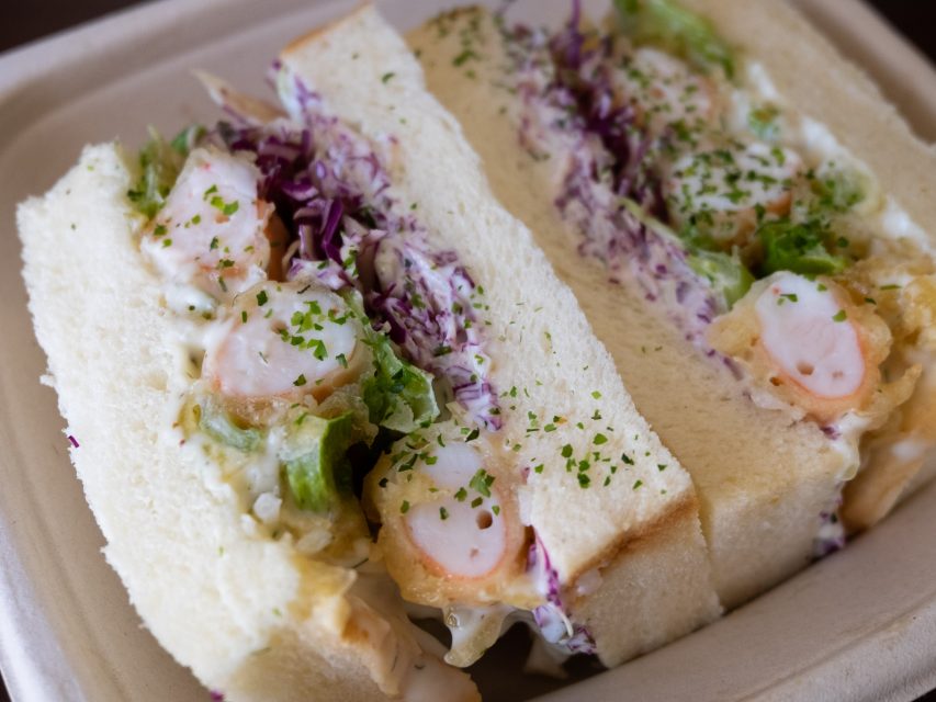 Close-up of a sandwich with shrimp, lettuce, red cabbage, and mayonnaise on soft white bread, garnished with chopped herbs.