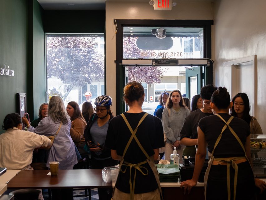 A bustling cafe with patrons standing in line and baristas working behind the counter. Several people are engaged in conversation, while others wait to place their orders. Trees are visible through the large window.