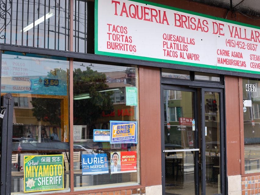 一家墨西哥餐廳的外觀，有標誌廣告各種食品如玉米餅和捲餅。政治競選海報展示在窗戶和門上。門上方可見一張「通過」證書。