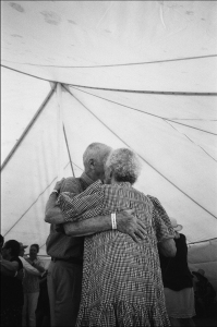 An elderly couple embraces and dances under a large tent at an event.