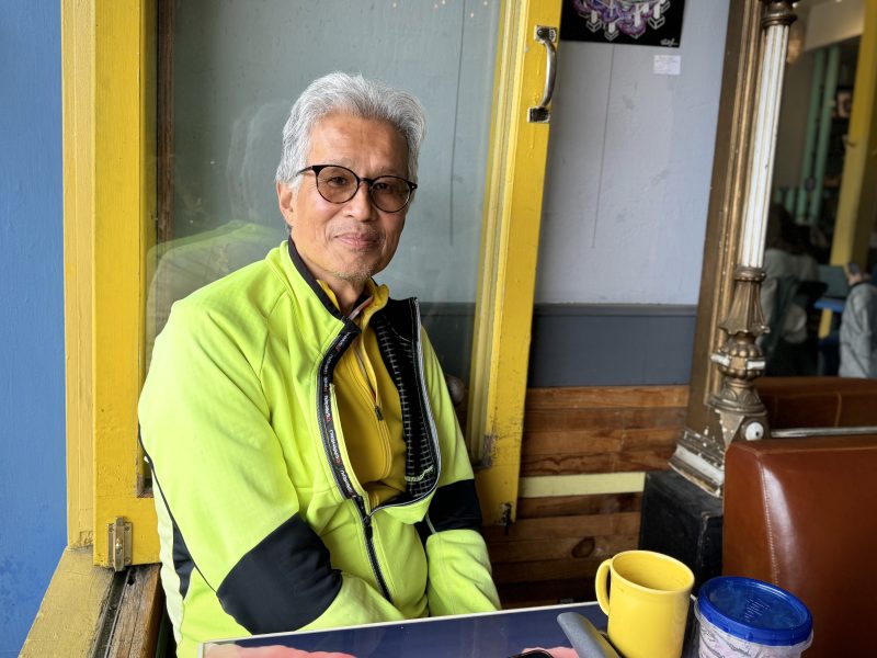 An older person with gray hair and glasses wearing a yellow jacket sits at a table with a blue Tupperware container and a yellow mug in front of him in a cafe.