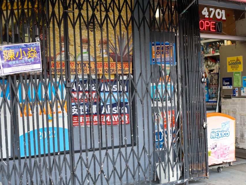 一家便利店入口，金屬安全門關閉。店面展示著各種標誌和廣告，包括貨幣兌換和彩票通知。商店位於 4701 號。