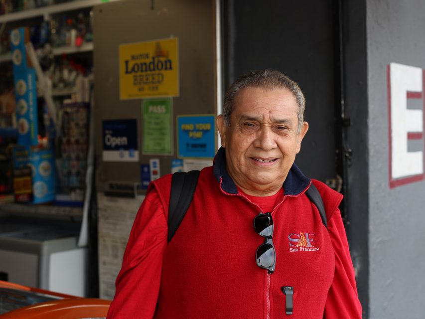 一位戴著眼鏡掛在脖子上、穿著紅色背心的男士站在店面前微笑，店面展示著各種標誌和物品。