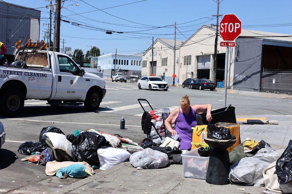 A person organizes belongings on a city sidewalk beside a street cleaning truck. Numerous bags and items are scattered around, ready to be swept up. The scene is set in an industrial area with buildings and power lines visible.