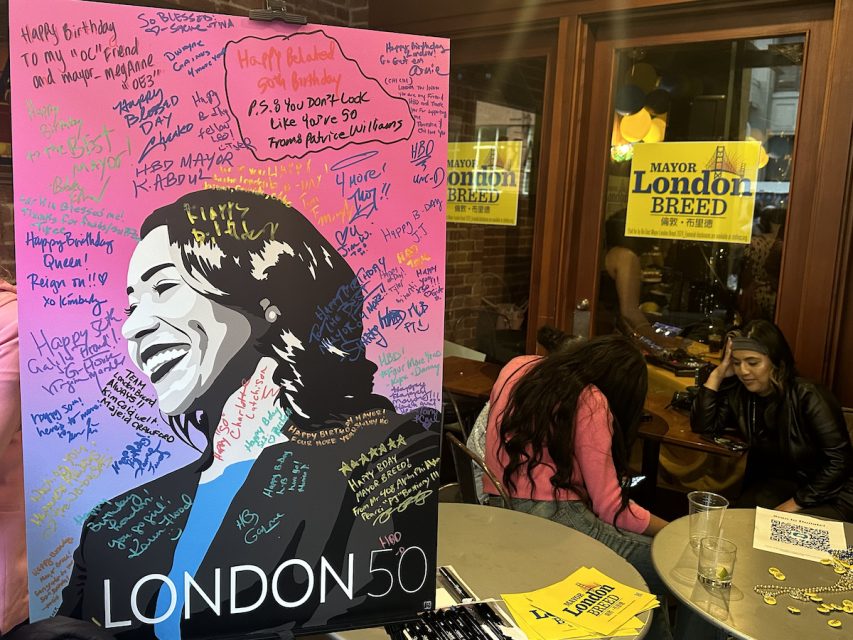 A large poster with a portrait of London Breed is covered with handwritten birthday messages. Two individuals are seated at a table in the background of a room with yellow banners reading "Mayor London Breed".