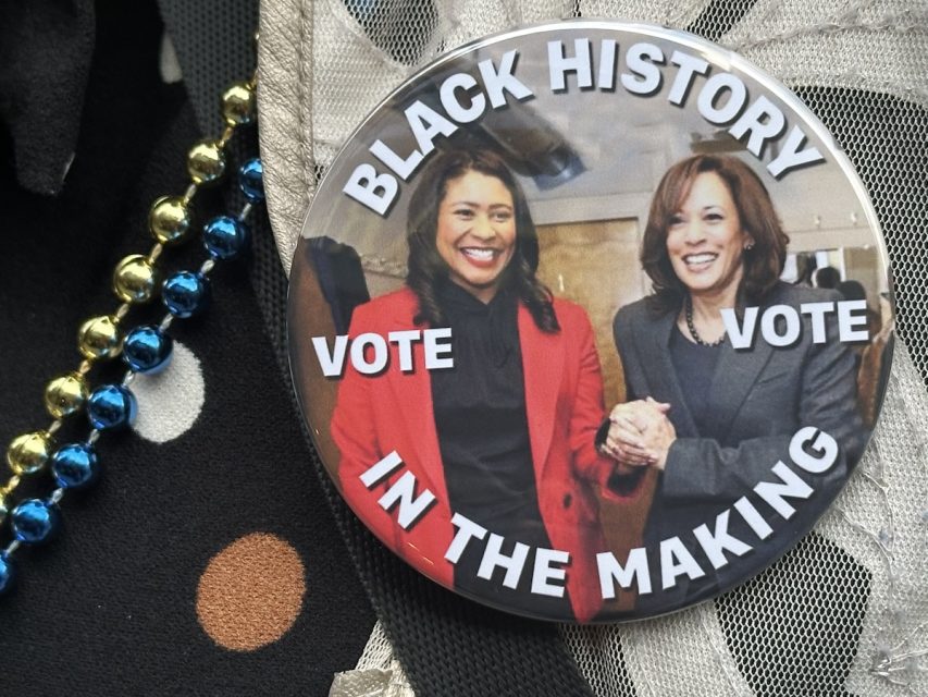 A button featuring two women smiling, with the text "BLACK HISTORY IN THE MAKING" and "VOTE" written around them. The button lies on a black fabric with polka dots and next to some metallic beads.