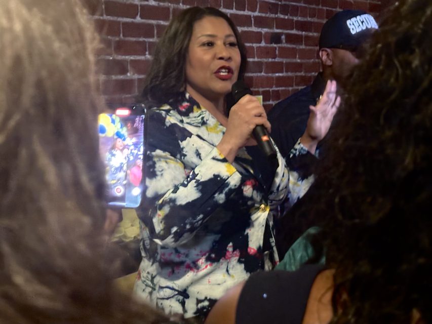 Mayor London Breed in a patterned jacket speaks into a microphone while standing in front of a brick wall. A crowd surrounds her, with one person holding up a phone to capture the moment.