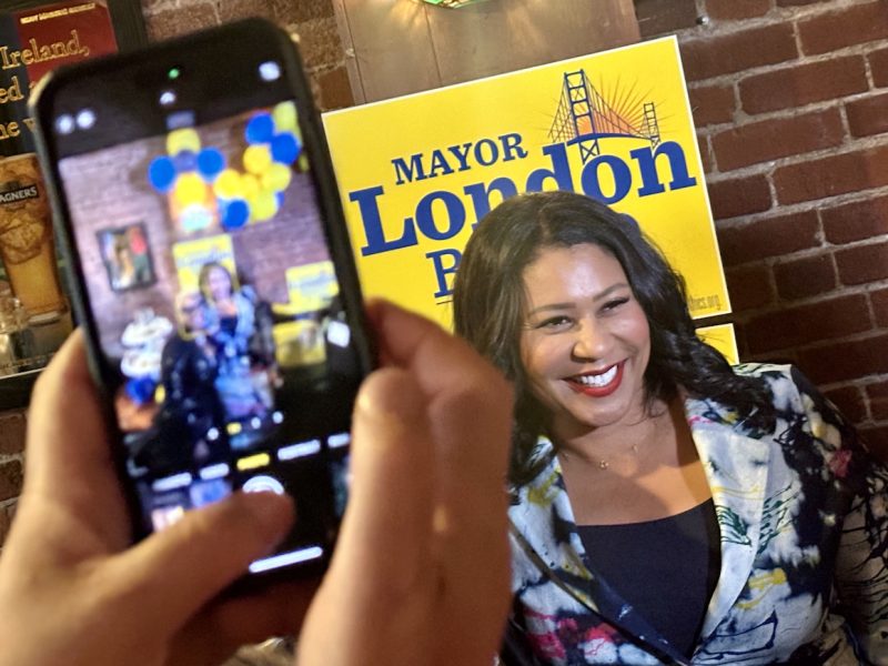 London Breed smiles for a photograph while being recorded on a smartphone. A campaign sign in the background reads "Mayor London Breed."