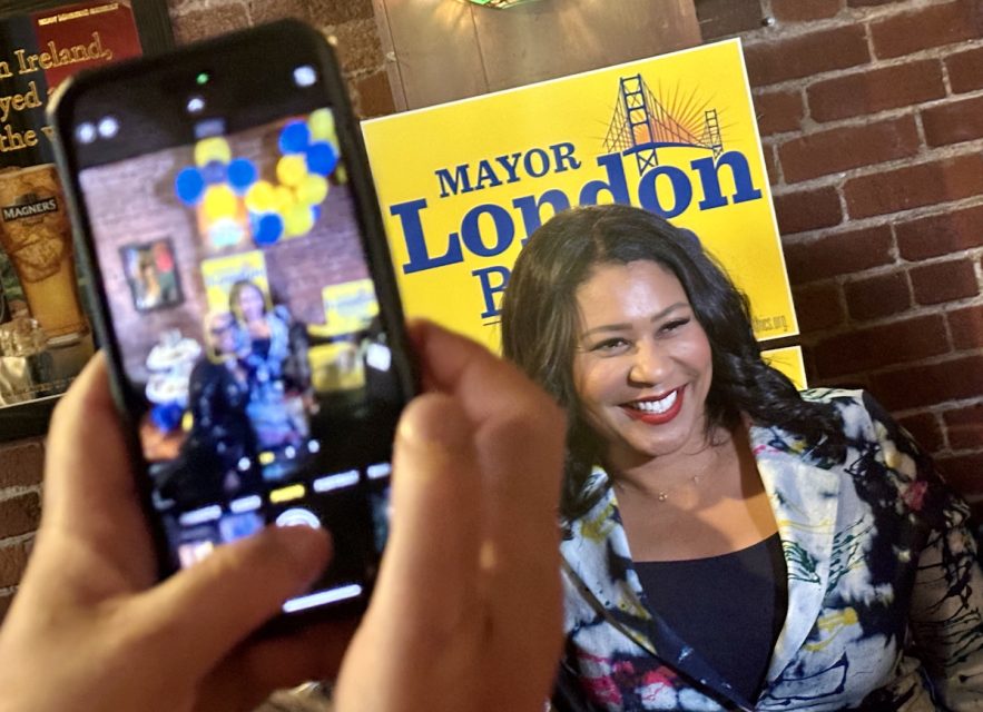 London Breed smiles for a photograph while being recorded on a smartphone. A campaign sign in the background reads "Mayor London Breed."