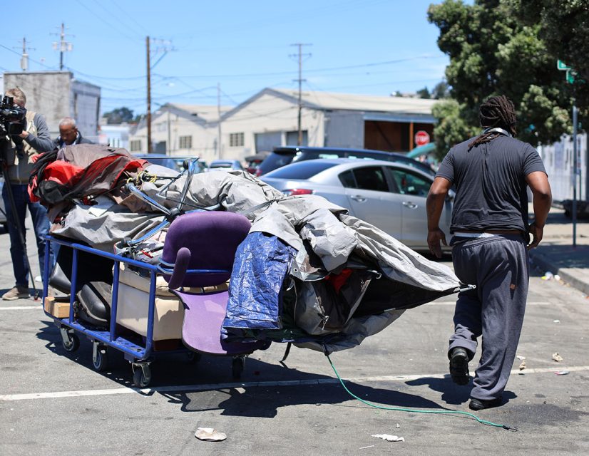 A man sweeps the street as he pulls a cart filled with various items, including a tarp and a purple chair.