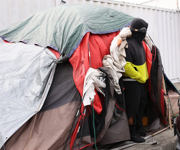 A person dressed in outdoor clothing stands partially inside a makeshift tent made of various colored tarps and fabric, situated next to a white container, as they begin to sweep the area around their temporary shelter.