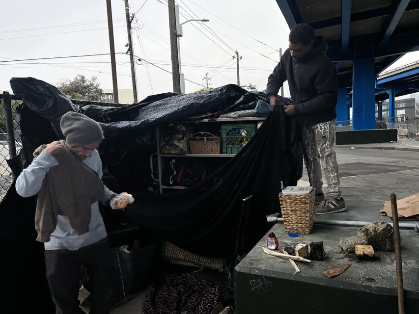Two people arrange a tarp and belongings under an overpass, with various personal items and makeshift furniture around them.