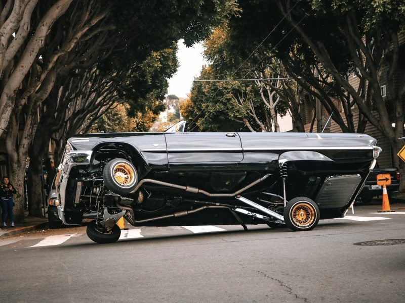 A lowrider car with hydraulics is tilted at an extreme angle on a tree-lined street, showcasing its modified suspension system.