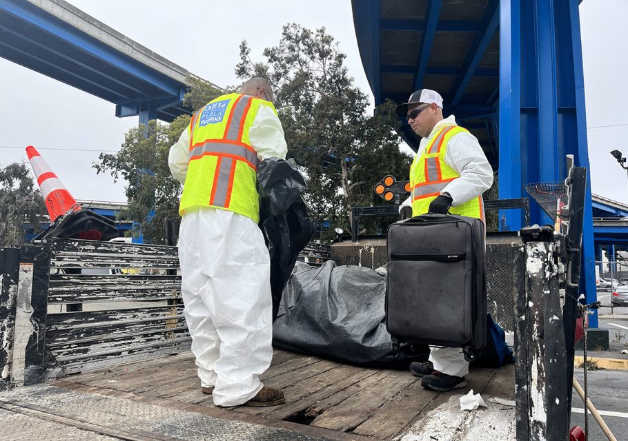 Two workers in white protective suits and yellow safety vests load items from a truck bed. One holds a black bag while the other carries a black suitcase. They are under a blue overpass.