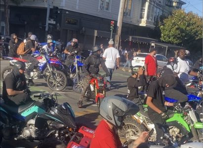 Group of people on motorcycles, some wearing helmets and masks, gathered at an urban intersection with buildings and pedestrians in the background.