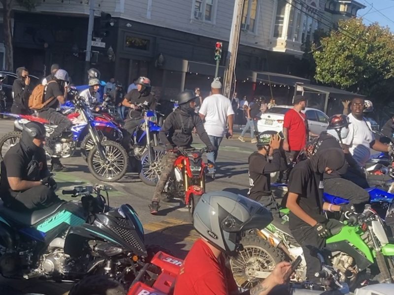 Group of people on motorcycles, some wearing helmets and masks, gathered at an urban intersection with buildings and pedestrians in the background.