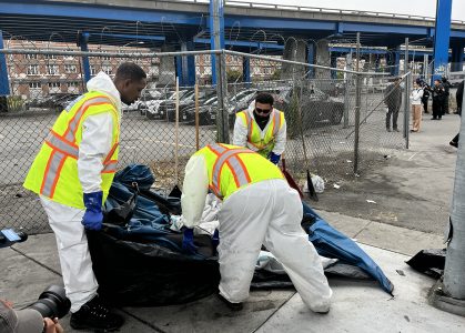 Three individuals wearing white hazmat suits and yellow vests clean up debris near a fenced area under an overpass. A person is visible in the foreground with a camera. Homeless sweep.