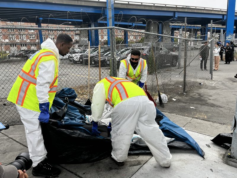 Three individuals wearing white hazmat suits and yellow vests clean up debris near a fenced area under an overpass. A person is visible in the foreground with a camera. Homeless sweep.