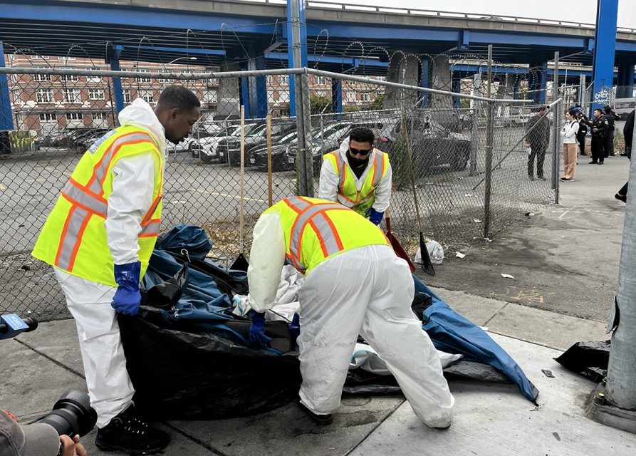 Three individuals wearing white hazmat suits and yellow vests clean up debris near a fenced area under an overpass. A person is visible in the foreground with a camera. Homeless sweep.