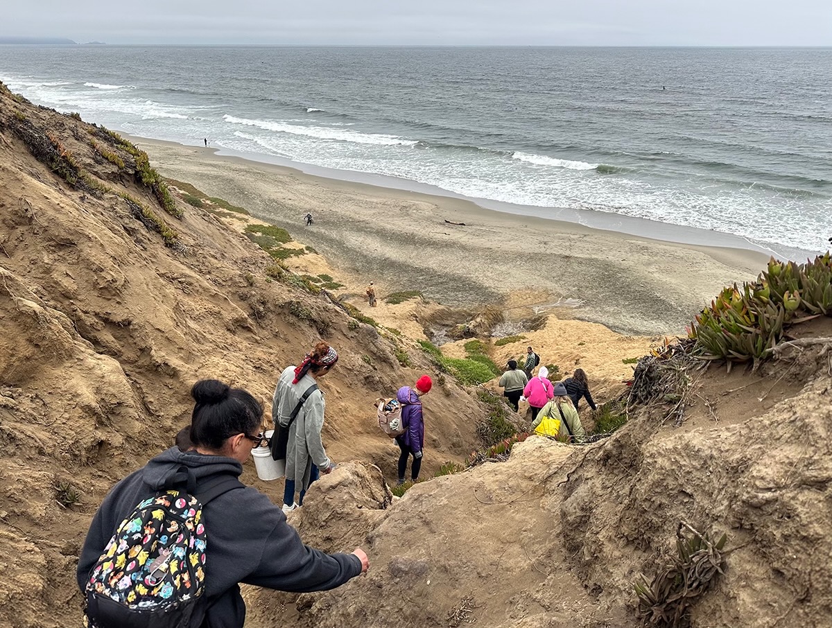 A group of people descends a rocky cliff path towards a sandy beach with waves gently washing ashore under a cloudy sky.