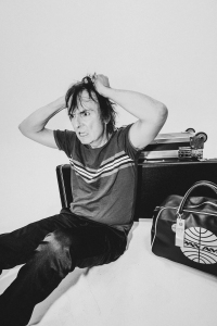 A person sits on the floor against a white wall, pulling their hair with both hands. A suitcase and a large bag are next to them.