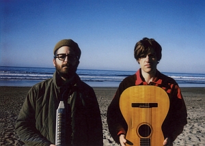 Two people stand on a beach with an ocean in the background. One holds a melodica, and the other holds an acoustic guitar.