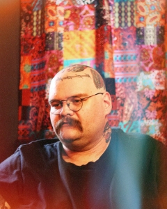 A person with a mustache, glasses, and tattoos on their head and neck sits against a colorful, patterned fabric backdrop.