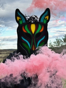 Person wearing a colorful animal mask with neon details, surrounded by pink smoke, in an outdoor setting with a cloudy sky.