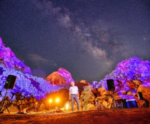 A person stands alone on rocky terrain illuminated by colorful lights, with a star-filled night sky in the background. Audio equipment is set up on either side.