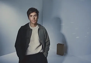 A person stands in a minimalist, softly lit room, wearing a light sweater and dark jacket, with hands in pockets. A cardboard box is placed on the floor near a wall in the background.