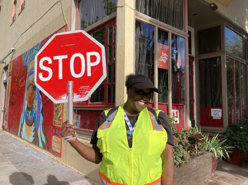 A person wearing a yellow safety vest and a black cap is holding a stop sign on a sidewalk in front of a building with murals.