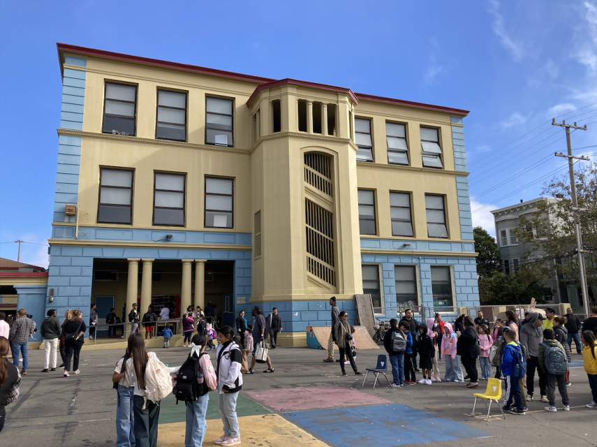 A three-story building with students gathered outside; some are standing while others are seated on plastic chairs. The building has blue and yellow sections with multiple windows and an arched entrance.