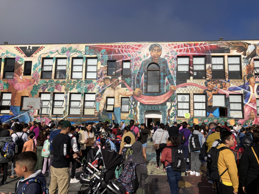 A large crowd of people, including many children, gathers in front of a colorful mural on a multi-story building. The mural features various figures and designs.