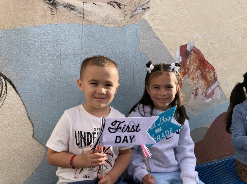 Two children are sitting, smiling, and holding signs that say "First Day." They are in front of a mural on a wall.