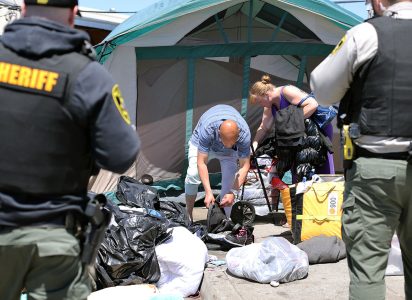 Sheriff officers oversee as individuals gather belongings in front of a tent during the sweep. Various items and bags are scattered on the ground.