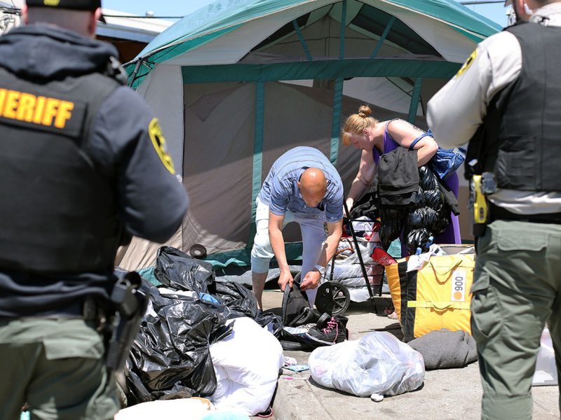 Sheriff officers oversee as individuals gather belongings in front of a tent during the sweep. Various items and bags are scattered on the ground.