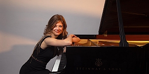 A woman in a black dress leans on the side of a black Steinway & Sons grand piano while smiling at the camera.