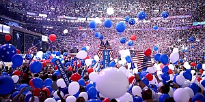 A large crowd is gathered at an indoor event, with red, white, and blue balloons and American flags visible. The atmosphere appears festive and celebratory.