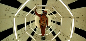 A person in an orange spacesuit is floating in a spacecraft's hexagonal corridor.