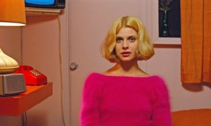 A woman with blonde hair wearing a bright pink sweater sits in a room with a red rotary phone, an orange curtain, and a vintage television.