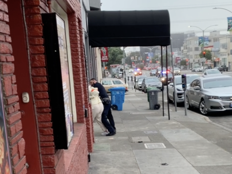 A person appears to be restrained by a police officer on a sidewalk. Several police cars with flashing lights are visible in the background on a city street.