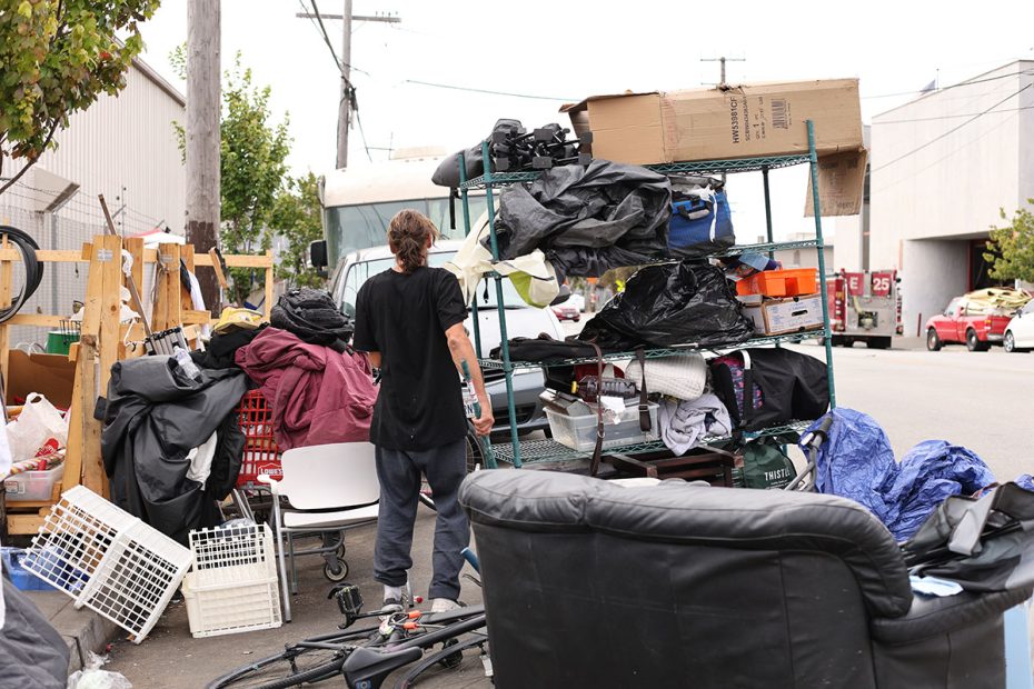 A person stands outdoors near a cluttered metal rack and an assortment of belongings, including boxes, furniture, and bags, in an urban environment. They appear poised to sweep through the disarray in an attempt to bring some order to the chaos.