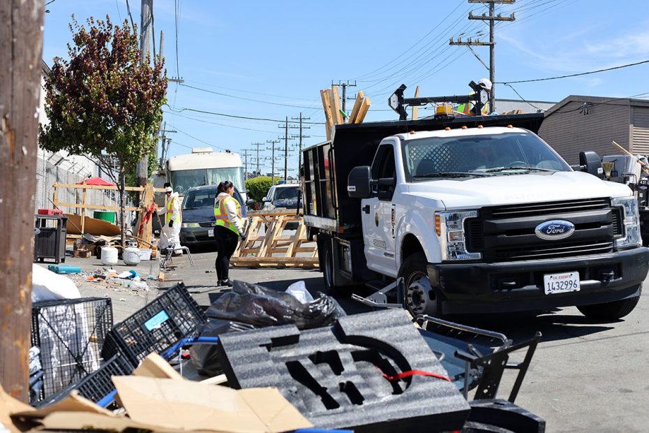 A cleanup crew in safety vests works near a white utility truck in an urban area, sweeping and clearing debris from the street, beside buildings and parked vehicles, under clear blue skies.