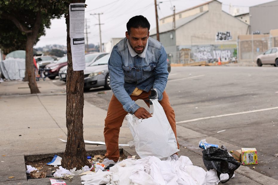 A man in a denim jacket and brown pants sweeps up items around a pile of white clothes and trash on a sidewalk.