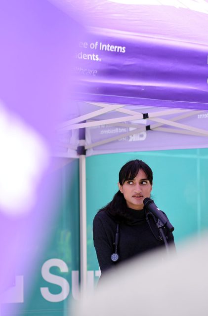 A person with long dark hair speaks into a microphone under a purple canopy.