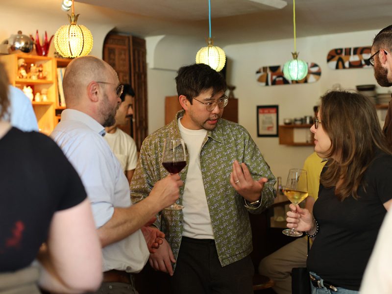 Several people are gathered in a room with hanging lights, holding wine glasses and engaged in conversation.