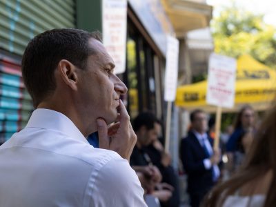 A man in a white shirt stands with his hand on his face, looking thoughtful. A group of people holding signs is visible in the background, suggesting a possible protest or gathering.