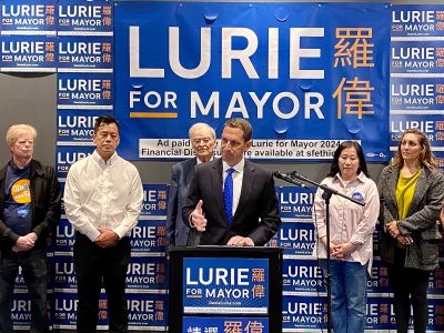 A mayoral candidate speaks at a podium during a campaign event, flanked by supporters. The backdrop features multiple "Lurie for Mayor" signs in English and Chinese.