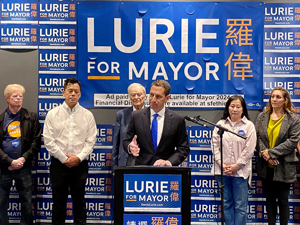 A mayoral candidate speaks at a podium during a campaign event, flanked by supporters. The backdrop features multiple "Lurie for Mayor" signs in English and Chinese.